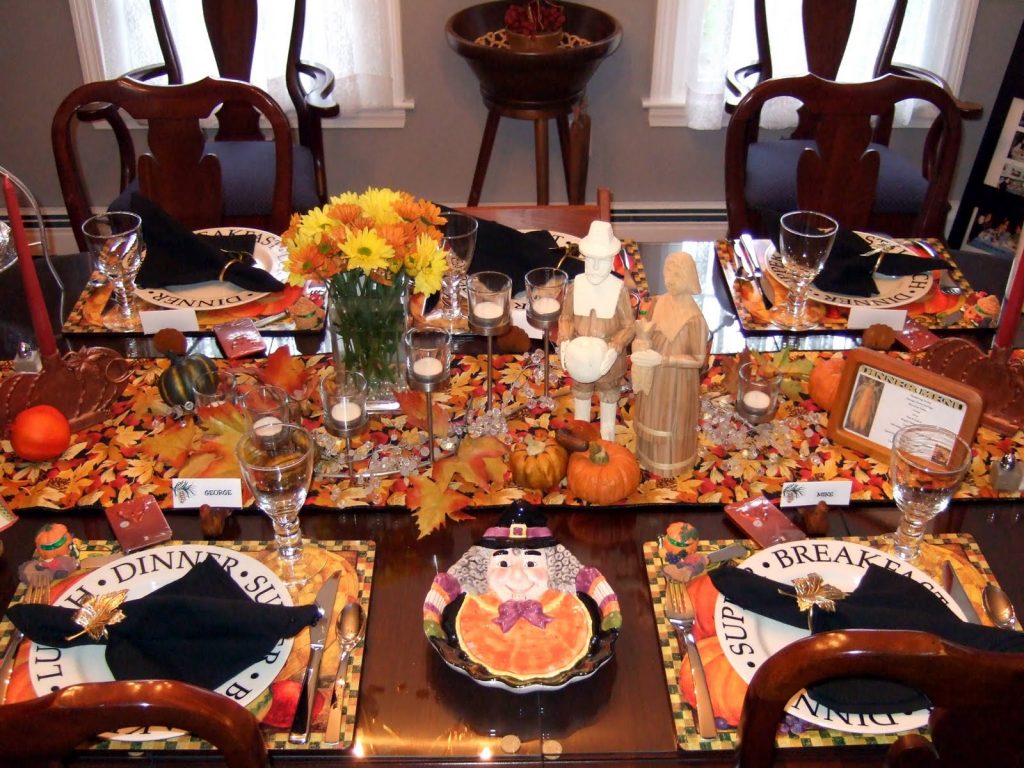 thanksgiving-tablescapes; a bright tavble runner scattered with leaves and mini pumpkins, a vase with orange and yellow flowers, and fun thanksgiving plates